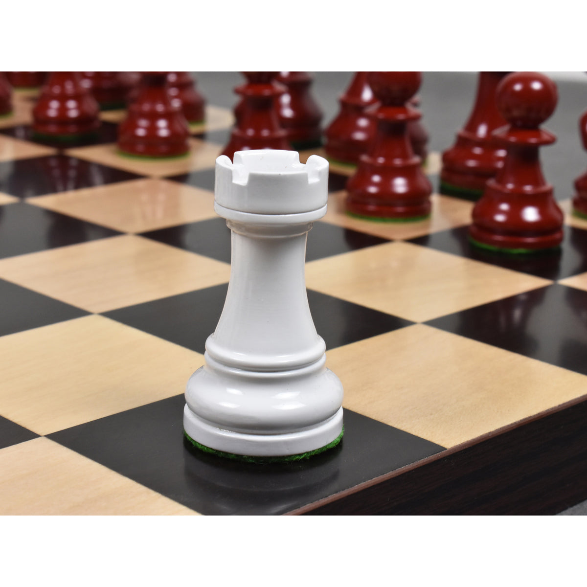Close-up of a white lacquered boxwood Prime Staunton rook on a chessboard, with red pieces softly blurred in the background.