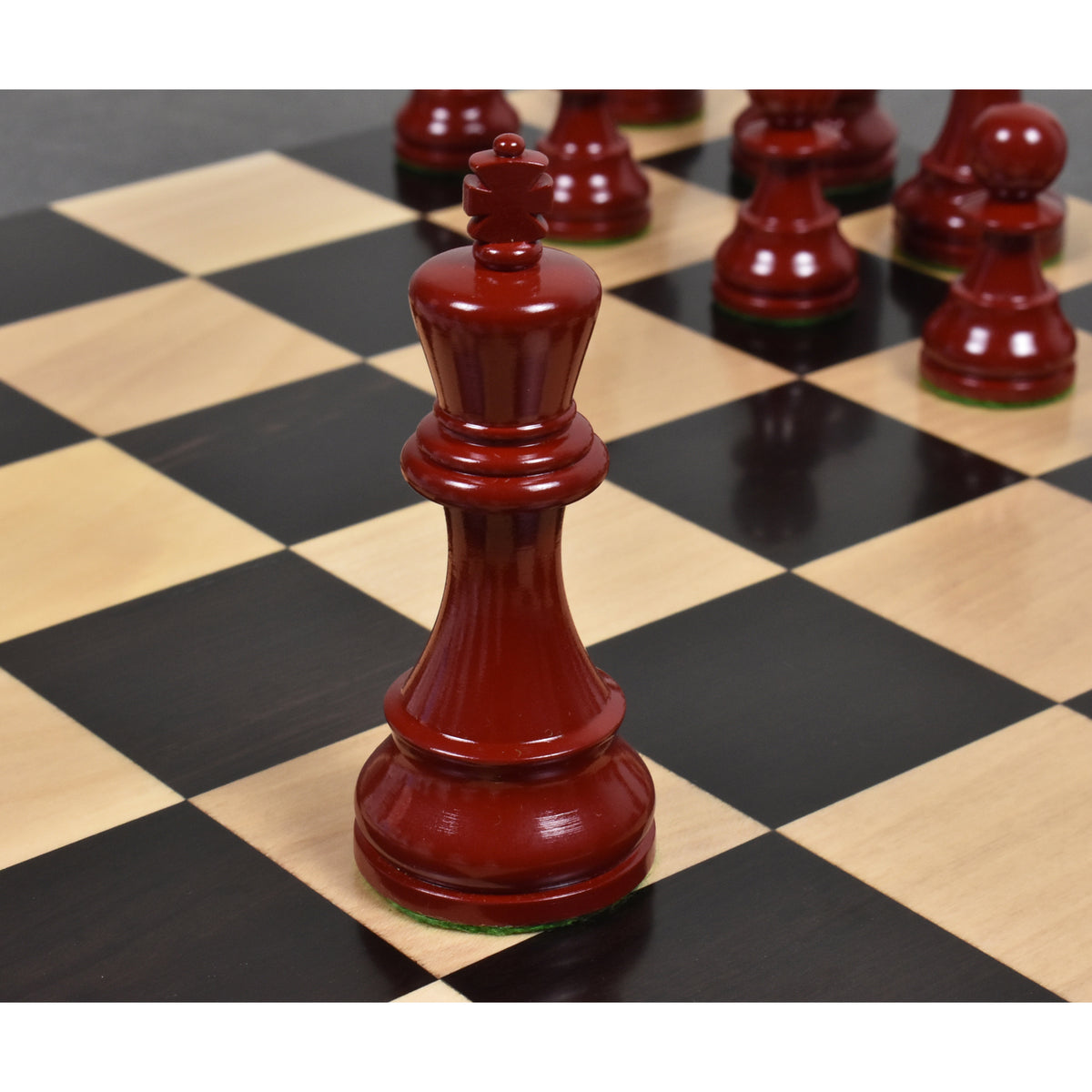 Close-up of a burgundy lacquered boxwood Prime Staunton king on a chessboard, with other red pieces blurred behind it.