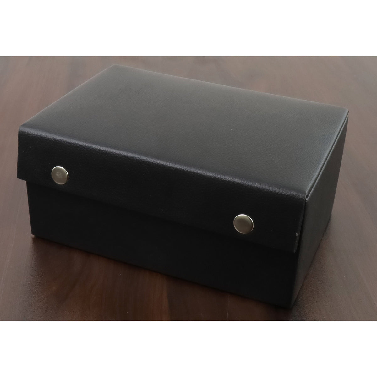 Closed black leatherette chess piece storage box with button fasteners on a wood surface.