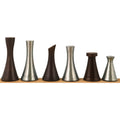 Six minimalist brass tower chess pieces in alternating dark brown and silver tones lined up on a wood base.