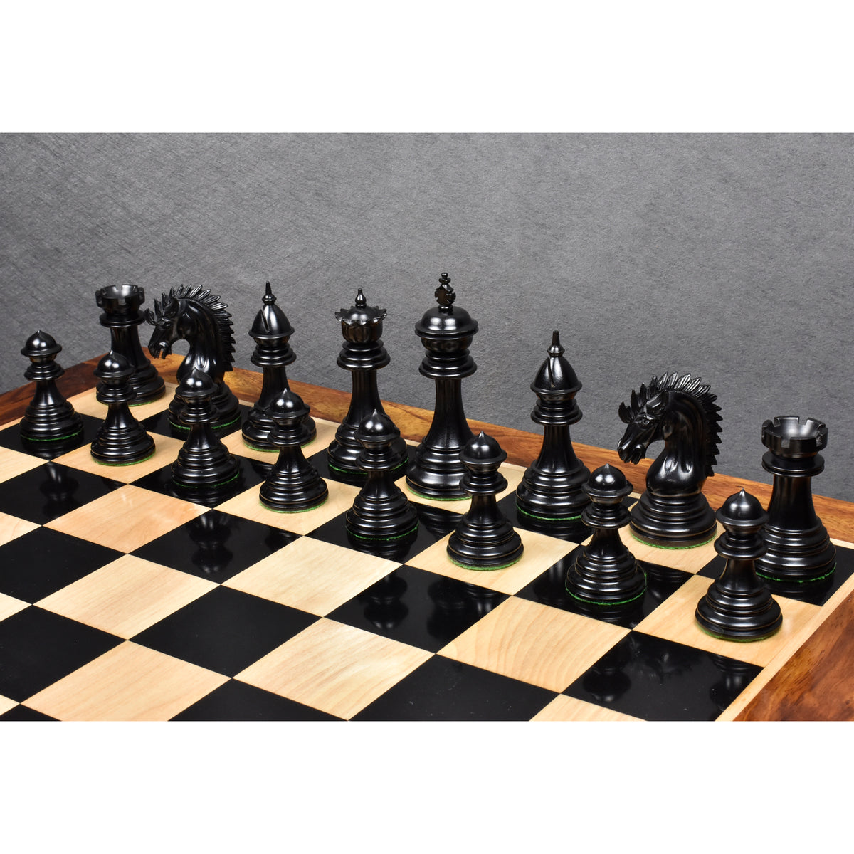 Polished dark ebony wood chess pieces set up along the edge of a glossy black and light maple chessboard.