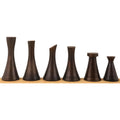 Six dark brown minimal brass tower chess pieces lined up in a row on a light-colored wooden stand.