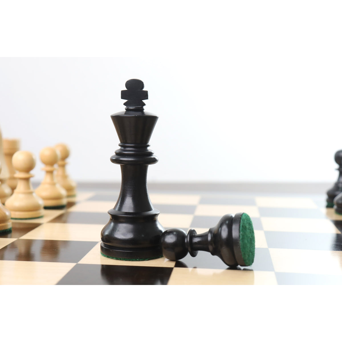 Ebonised boxwood king piece standing on a tournament chessboard next to a toppled black pawn.